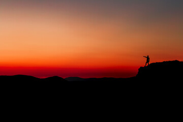 Red sunset with mountain silhouette. A man on mountain in sunset. Traveler on rock waving hands.