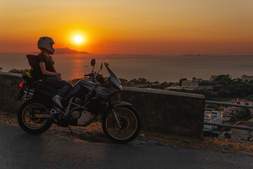 Fototapeta na wymiar Biker girl sits on a adventure motorcycle. Freedom lifestyle concept. Romantic sunset. Sea and mountains, Copy space. Capri island. Sorrento Italy