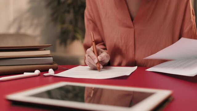 Steadicam of hands of cropped unrecognizable person wearing peachy blouse sitting at table, proofreading and making notes on papers