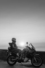 Fototapeta na wymiar Biker girl sits on a adventure motorcycle. Freedom lifestyle concept. Romantic sunset. Sea and mountains, Black and white. Vertical photo. Capri island. Sorrento Italy