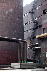 Vintage Soviet industrial building with stairs and broken windows, smoke stack (left) and metal pipes in rain or snow. 