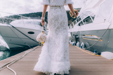 A beautiful bride in a vintage dress with a wedding bouquet in her hand walks along the sea pier near the yachts. - 427976653