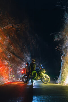 Motorcyclist biker man with a motorcycle for long journeys. A road in a narrow canyon. Night and lights. Tourism and vacation. Vertical photo.