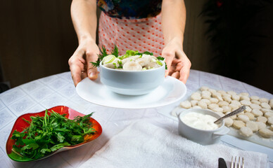 Hands of a woman in an apron hold out a bowl of cooked homemade dumplings, decorated with green leaves of dill and parsley. Selective focus.