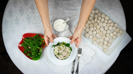 Hands of a woman hold out a bowl of cooked homemade dumplings, decorated with green leaves of dill and parsley against background of white round table. Flat lay.