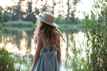 Cute girl in a blue dress and straw hat stands on the shore of the lake. Back of a child in the public park with trees, green and dried grass in summer. Young beautiful lady with curly hair on walking