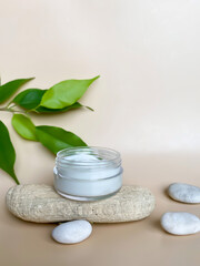 Spa background for natural cosmetics.  A jar of cream on a podium made of natural natural material, wood, sea pebbles and green leaves on a beige background.  Copy space.