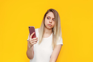 Naklejka premium A frightened young caucasian blonde woman in a white t-shirt with a mobile phone in her hand looks suspiciously at the incoming call from an unknown number on a bright color yellow background. Fraud