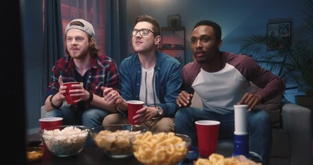 Mixed-race young handsome males sitting on sofa in living room at home at night watching sport channel on TV feeling sad disappointed when team lose game match. Entertainment, leisure concept