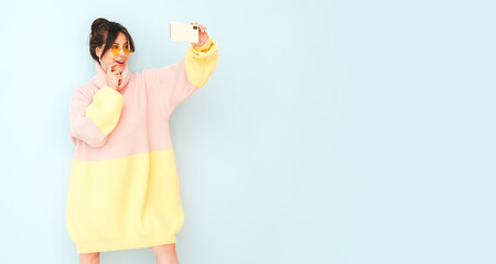 Young beautiful smiling female in trendy summer colorful sweater. Sexy carefree woman posing near light blue wall in studio. Positive model having fun indoors in sunglasses.Taking selfie