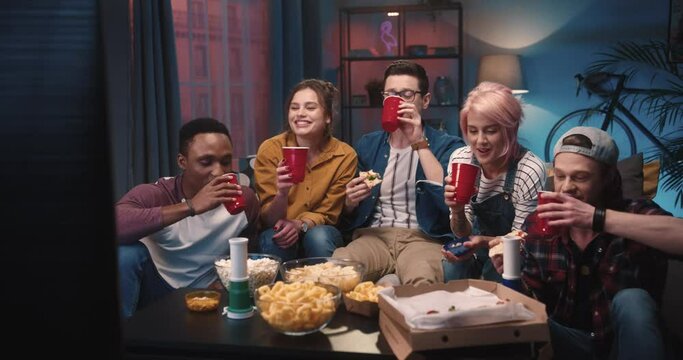 Happy mixed-race young company sitting at home in living room at night gathered together drinking having fun. Friends having drinks while watching sport game in house Sport fans, entertainment concept