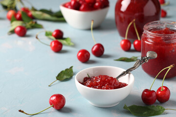 Homemade jam of sweet cherry in a white bowl and jars on light blue background. Close-up