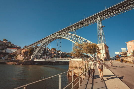 People walk past riverside under the old bridge on bright sunny day. Porto.