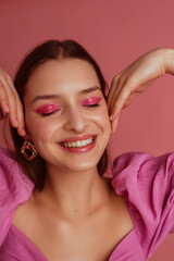 Happy smiling beautiful woman with pink, fuchsia color eyeshadow makeup, flawless clean skin, wearing elegant golden earrings, pink blouse. Spring, summer fashion trends
 - obrazy, fototapety, plakaty