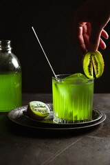 A rocks glass with a refreshing green drink, a bar spoon and an ice cube inside, garnished with kiwi, a bottle of drink in the background. Hand holding kiwi garnish. Dark backdrop, vertical photo.