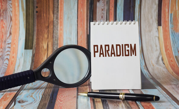 The word paradigm is written on a white notebook, near a pen and a magnifying glass on a striped background