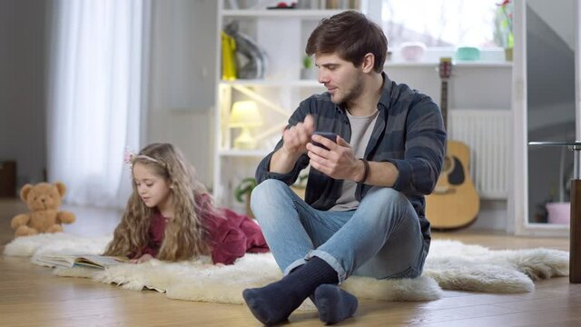Absorbed handsome young man messaging online on smartphone distracted by cute little girl drawing picture. Portrait of busy Caucasian father ignoring pretty daughter at home indoors