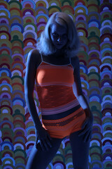 Eine Frau steht im Dunkel gekleidet in orangenen Dessous vor buntem Hintergrund