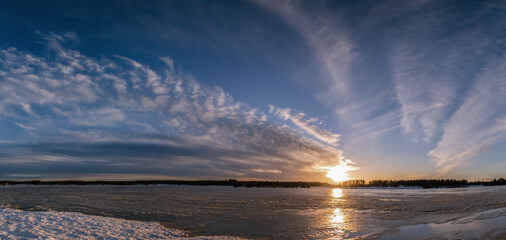 Scenic wide panorama of spring sunset over melting icy lake. Beautiful red yellow orange Sun, dark...