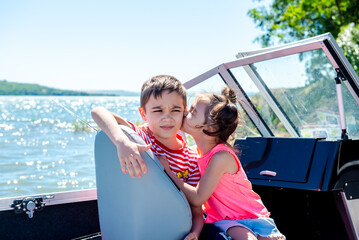 Fototapeta na wymiar Happy boy and little girl are sitting in a motor boat on the shore of a lake or river. Summer vacation. Rest with children by the water. Brother and sister 