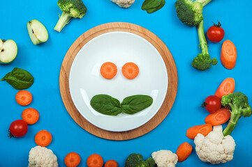 Plate and smiley food on blue background surrounded by fruits and vegetables. Complementary feeding for children, food allergy, diet, healthy eating concept. Spinach, broccoli, cauliflower, carrot, ap