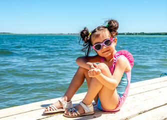 A cute little girl is sitting on a wooden pier. The kid is resting by the sea. Summer vacation. A...