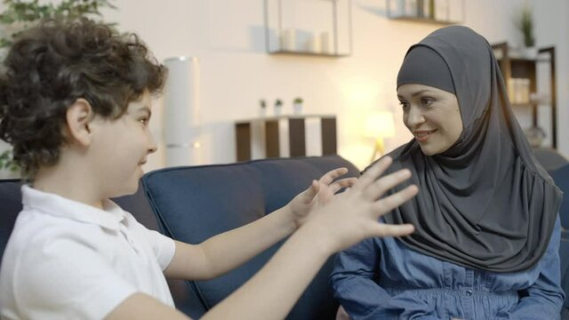 Cute arabic boy telling mom funny story, happy woman in hijab laughing, family