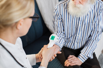 Close up cropped shot of female doctor checking body temperature of senior bearded man patient. Body temperature measurement with infrared wrist non-contact thermometer