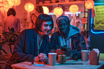 Young man sitting at the table with his boyfriend they watching something on mobile phone sitting in nightclub