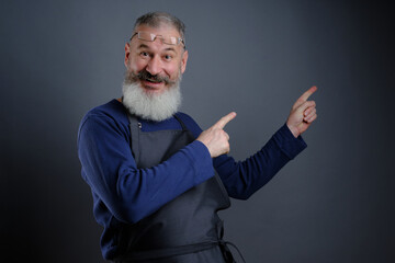 Follow me. Look here. Close up portrait of cheerful pleased qualified experienced joyful apron-wearing grandfather pointing on empty blank place over shoulder isolated on grey background