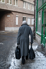 Old lady with gray coat and a plastic bag on her head.
She lady walks on the snowy sidewalk holding two black bags.