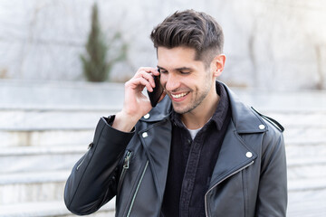 Close up portrait of a handsome young man talking on the phone with a smile in a leather jacket outdoors, modern businessman 