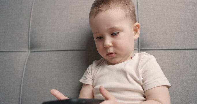 Baby boy sits on the bed holding a black phone and looking at it. The concept of using gadgets by children. Close-up, slow motion