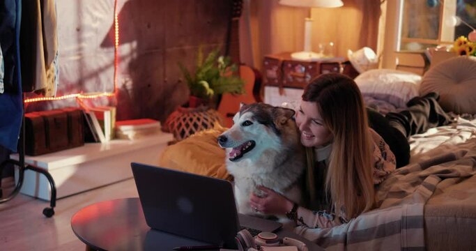 Happy smiling girl and her dog Husky look at a laptop. Young woman hugs and strokes dog. Girl and a dog watch a movie late at night