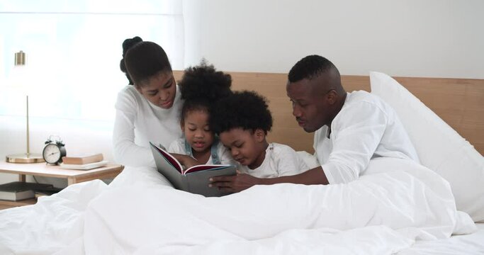 Happy African American dad and mom reading a book with their adorable children in bedroom. little cute kids listens fairy tale story on bed with their parent. Development for kids concept.