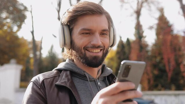 Portrait of Smiling Bearded Guy watching Video on Smartphone through Headphones. Adult Man Browsing in Social Media while Standing on the Street. Wearing casual jacket on Spring Weather. Mobile.