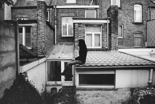 Portrait of girl sitting on a roof.
Brunette girl dressed in black surrounded by windows and bricks.
Black and white photo and architecture.