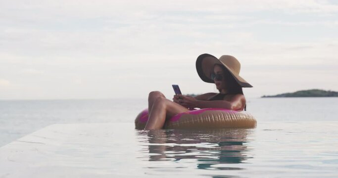Vacation woman on inflatable donut float mattress using mobile cell phone in swimming pool. Girl relaxing relaxing enjoying travel holidays at resort infinity pool in bikini. Luxury lifestyle.