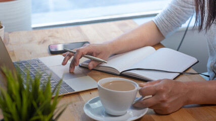 Obraz premium Woman working on laptop in office. Close up of desk, with coffee cup, notepad, and laptop. Holding a pen.