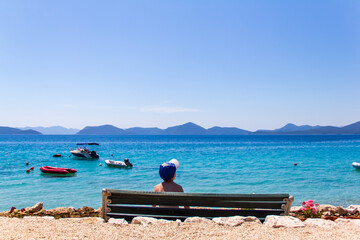 Kid sitting on bench at the beach and starring at the blue cristal sea during summer sunny day. Boats floating over the see. in the background mountains and islands. Slano, Croatia.