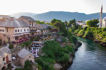 Fototapeta na wymiar View from the top of Old Bridge in Mostar, Bosnia-Herzegovina during summer sunny day. Bridge over the river and very nice shores with small restaurants and building with the mountains in the back.