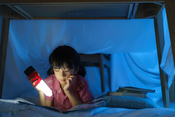 Asian girl child to make a camp to play imaginatively and reading book by flashlight  in living room at home..