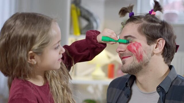 Side view close-up of cheerful pretty girl painting father eyelids with green marker smiling. Positive Caucasian daughter applying makeup on parent face at home on weekend. Family leisure and beauty