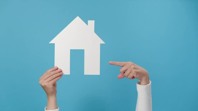 Close up of young female hand raising and pointing to small white paper house, posing isolated on blue studio background wall with copy space for advertisement. Property and mortgage concept