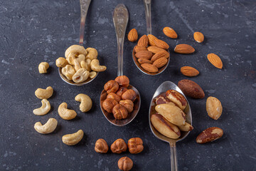 Assortment of various types of nuts: cashew, hazelnuts, almonds, brazil nuts on metal silver spoons on dark background. Healthy vegetarian snacks. Protein-containing food. Copy space for text