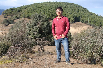 A Nepali Boy is wearing a brown color clothe and blue jeans and taking photo in front of forest