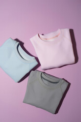 Flat lay of three folded pastel pink, blue and gray sweatshirts isolated on pale purple background