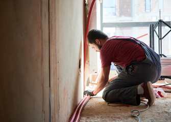 Young plumber connecting water radiator to heating system in new unfinished apartment. Crouching man working with hammer. Building process. Construction, maintenance concept