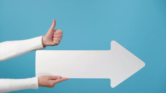 Close up of unrecognizable young female showing aside, pointing and holds white paper arrow with thumbs up like gesture, isolated on blue studio background wall with copy space for advertisement