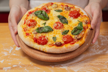 Chef holds wooden tray or board with homemade cheese pizza with tomato cherry, sauce, sausage and basil. Tasty dinner, close up view